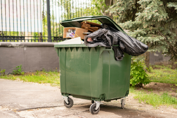 A full, dark green dumpster on wheels sits outside in an urban setting with a tall fence behind it and greenery growing around the edges. The dumpster is full of trash including a black trash bag. The dumpster could be used in a variety of projects related to trash collection, environmental concerns, or urban settings. The photo has a gritty feel and could also be used in projects related to urban decay or neglect.