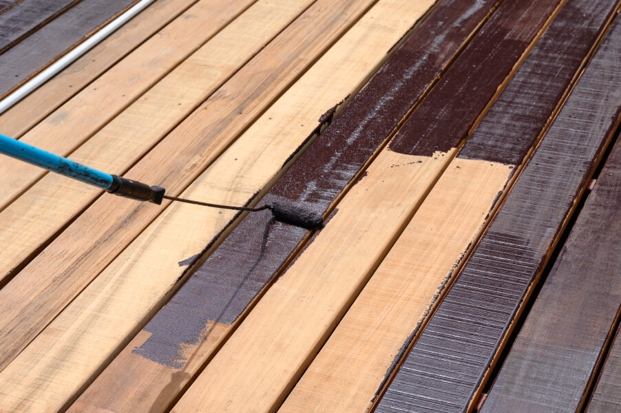 A paint roller with a blue handle applies dark brown stain to light-colored wooden boards.