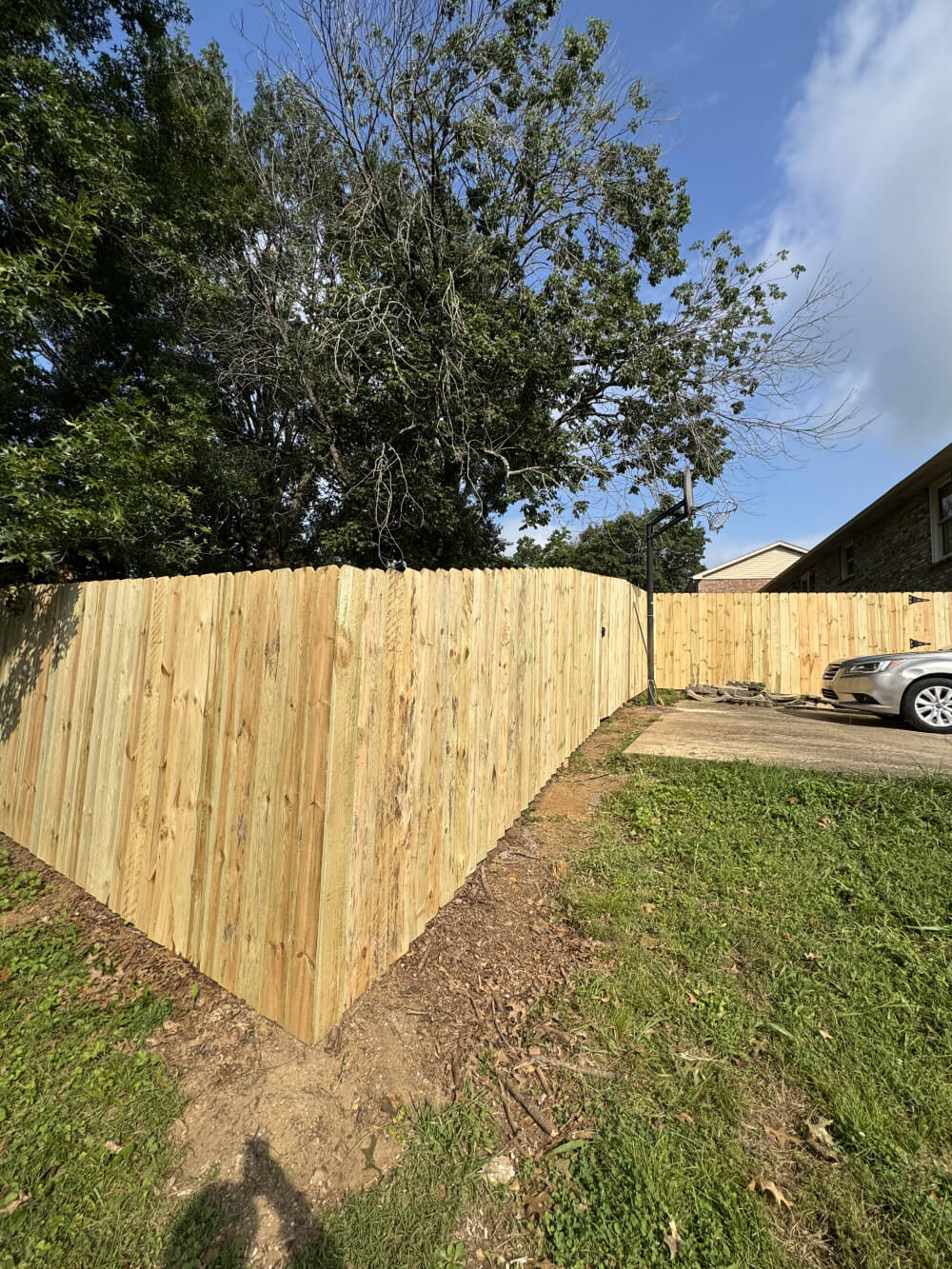 A tall wooden privacy fence making a corner bend along a grassy yard next to a paved driveway and basketball hoop.