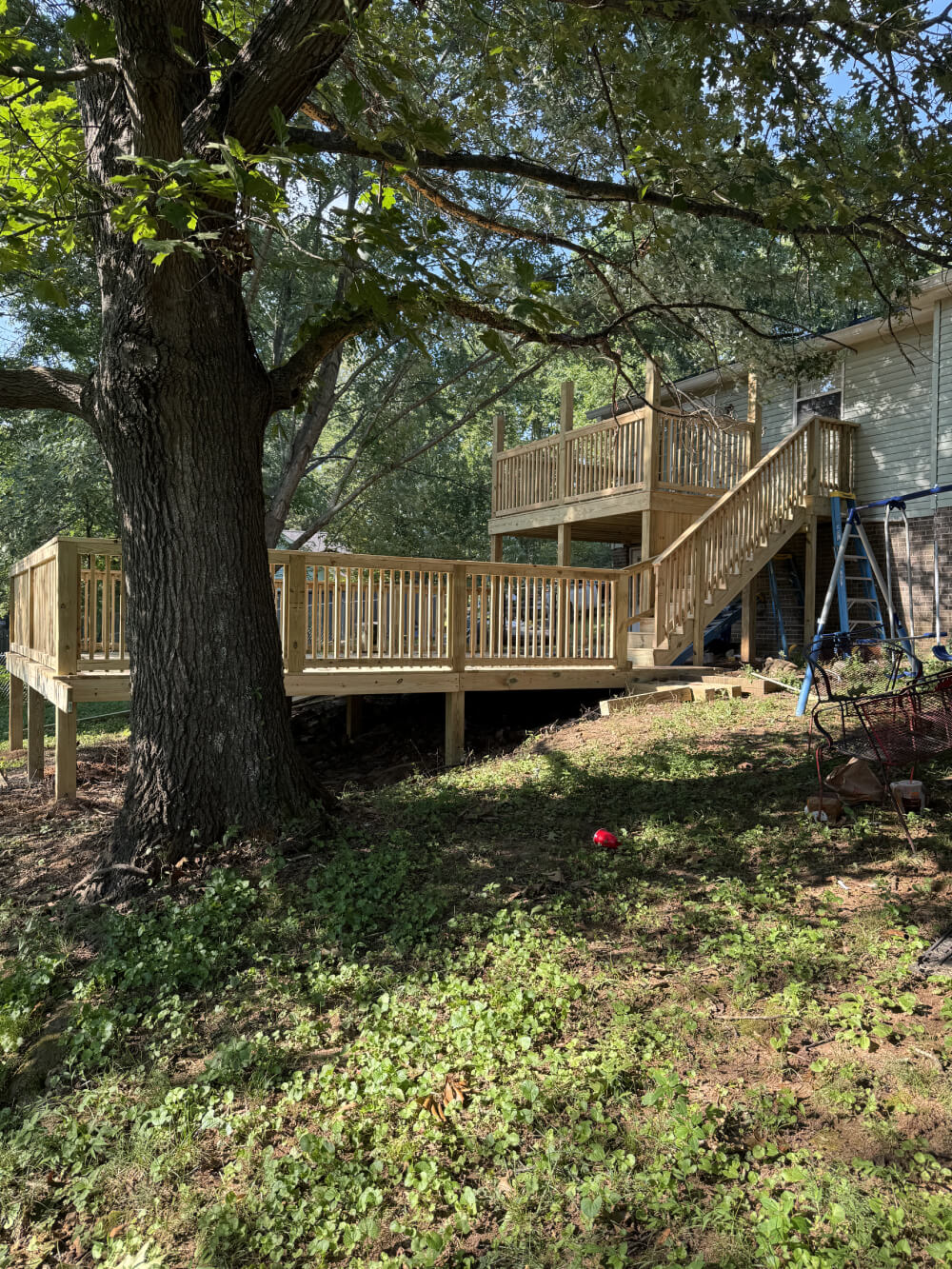 A newly built, multi-level wooden deck attached to the back of a house, surrounded by a grassy yard and large shady tree.